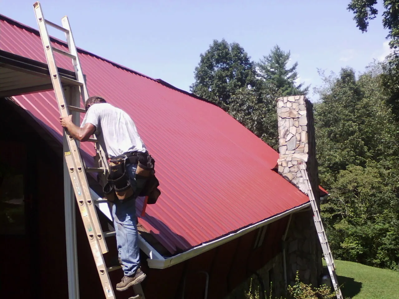 Roofer on a ladder beside a red metal roof and stone chimney — Soffit & Fascia Repair by Roofing Brothers in Occoquan